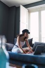 Приваблива молода стильна жінка в кепці використовує ноутбук, відпочиваючи на дивані біля балкона вдома — стокове фото