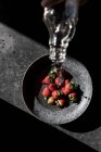 Abgeschnittene Hand einer Person, die eine Flasche hält und Wasser aus frischen Erdbeeren in eine Schüssel auf dunklem Hintergrund gießt — Stockfoto