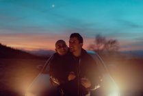 Glückliches homosexuelles Paar umarmt sich auf Wiese neben Auto mit beleuchteten Scheinwerfern am Abend — Stockfoto