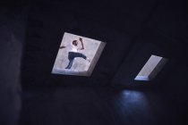 From below young man in casual outfit jumping over hole in roof of grungy building while dancing modern dance on street — Stock Photo
