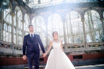 Знизу молодий елегантний чоловік тримає на руках жінку у весільній сукні біля ретро-палацу з багатьма вікнами в сонячний день — стокове фото