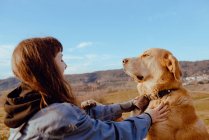 Вид сбоку на молодого хипстера, гладящего смешную собаку между лугом и голубым небом — стоковое фото