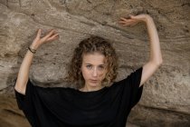Jeune femme blonde avec les mains levées regardant la caméra sous le rocher — Photo de stock