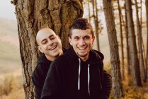 Fröhliches homosexuelles Paar, das sich umarmt und in die Kamera schaut, in der Nähe eines Baumes im Wald und malerischer Blick auf das Tal — Stockfoto