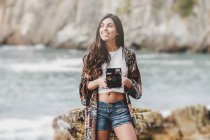 Smiling young woman holding camera on sea coast — Stock Photo