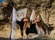 Jeunes ballerines mystérieuses en tenue noire avec des textiles blancs dansant près des rochers par temps ensoleillé — Photo de stock