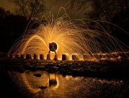 Long exposure of silhouette of human with burning round and fireworks on rocks near water in evening — Stock Photo
