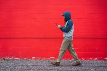 Vista lateral de um cara com barba trançada em um smartphone de navegação com capuz enquanto caminha perto da parede vermelha na rua da cidade — Fotografia de Stock