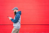 Вид збоку привабливого хлопця з плетеною бородою переглядає смартфон під час ходьби біля червоної стіни на міській вулиці — стокове фото