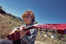 Cute boy playing acoustic guitar and singing while sitting in huge roll of dry hay on sunny day in countryside — Stock Photo