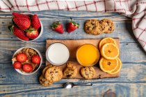Dall'alto occhiali di latte e succo vicino a cucchiaio, biscotti saporiti, fette di arancione e fragole su sfondo di legno — Foto stock