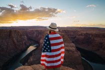Обратный вид человека, обернутого флагом США, стоящего рядом с красивым каньоном на фоне закатного неба на западном побережье — стоковое фото
