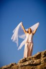 Снизу молодая загадочная женщина с поднятыми руками держит белый текстиль и позирует на скалах и голубом небе — стоковое фото