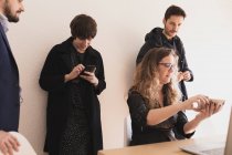 Jovens do sexo masculino e senhora conversando, navegando no celular e digitando no laptop à mesa no escritório — Fotografia de Stock