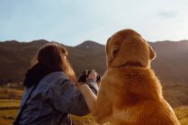 Vista posterior de la joven mujer disparando paisaje en la cámara cerca de perro divertido y amable entre el prado y el cielo azul - foto de stock
