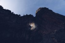 From below of huge dark rocky cliff with person climbing in spot of light under blue sky — Stock Photo