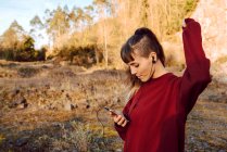 Joven mujer hipster con la mano levantada escuchando música con teléfono móvil y bailando en el campo - foto de stock