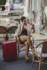 Femme élégante afro-américaine joyeuse tenant un téléphone portable et assise à table avec un verre de jus près des bagages dans un café de rue — Photo de stock