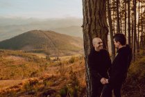 Cheerful homosexual couple looking at each other near tree in forest and picturesque view of valley — Stock Photo