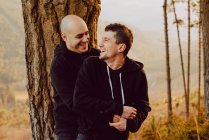 Alegre casal homossexual abraçando e olhando para a câmera perto da árvore na floresta e vista pitoresca do vale — Fotografia de Stock