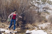 Side view of casual woman carrying case and jumping on rocks of clear stream of water in nature — Stock Photo