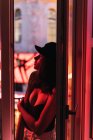Joven mujer delgada en la tapa de pie cerca de balcón en la habitación en el enrojecimiento por la noche - foto de stock
