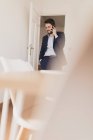 Концентрированный молодой мужчина с рукой в кармане разговаривает по мобильному телефону в комнате со стульями и столом — стоковое фото