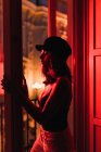 Молода струнка жінка в кепці, що стоїть біля балкона в кімнаті в почервоніння вночі — стокове фото