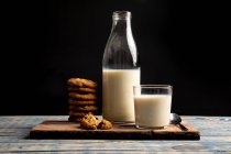 Vaso y botella de leche y pila de galletas frescas sobre tabla de madera sobre fondo negro - foto de stock