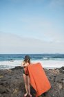 Slim hembra en traje de baño y sombrero que pone la toalla en la costa pedregosa cerca del mar ondulante en la magnífica naturaleza - foto de stock