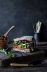 Sandwich of pate of dry tomatoes, fresh salad and cabbage on tray near knife on wooden board on black background — Stock Photo