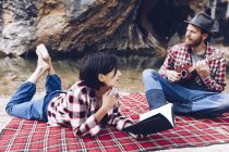 Hombre y mujer adultos en cuadros a cuadros con libro y pequeño ukelele teniendo picnic en la orilla del lago - foto de stock