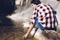 Вид ззаду жінка сидить на скелі і торкається чистої води в озері — стокове фото