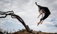 Dal basso giovane ballerina in abito nero con le mani in alto in aria vicino rami secchi e cielo blu nelle nuvole — Foto stock