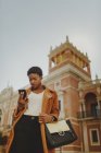 Mujer elegante afroamericana con confianza en la chaqueta que sostiene el bolso y el teléfono móvil en la calle - foto de stock