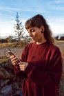 Joven mujer hipster con piercing y auriculares escuchando música con teléfono móvil y de pie en el camino del campo - foto de stock