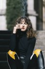 Привлекательная молодая женщина в стильном наряде сидит на лестнице и курит сигарету в солнечный день на городской улице — стоковое фото