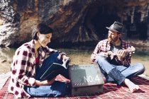 Uomo e donna adulti a scacchi plaid con libro e piccolo ukulele fare picnic sulla riva del lago — Foto stock