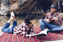 Erwachsener Mann und Frau im karierten Karo mit Buch und kleiner Ukulele beim Picknick am Seeufer — Stockfoto