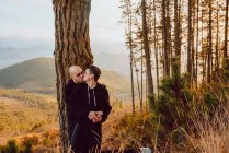 Romântico casal homossexual beijando e abraçando perto de árvore na floresta e vista pitoresca do vale — Fotografia de Stock