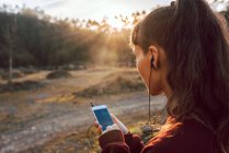 Молода хіпстерка з пірсингом та навушниками слухає музику з мобільним телефоном у сільській місцевості — стокове фото