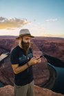 Привабливий бородатий чоловік у капелюсі на мобільному телефоні, стоячи проти чудового каньйону та річки під час заходу сонця на західному узбережжі Уси. — стокове фото