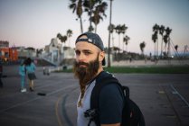 Side view of bearded guy with backpack looking at camera while standing on pavement on street of modern city — Stock Photo