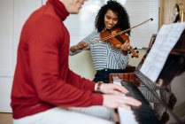 Погляд на молодого чоловіка, який грає на піаніно поруч з чорношкірою жінкою, що грає на скрипці в музичній студії — стокове фото