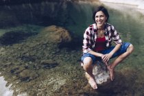 Glückliche erwachsene Frau sitzt auf einem Felsen im ruhigen, transparenten Wasser des Sees und genießt die Natur und lächelt weg — Stockfoto