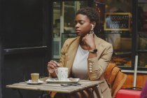 Mujer elegante afroamericana con auriculares mirando a la cámara, sosteniendo taza de bebida y sentada en la mesa en la cafetería de la calle sobre fondo borroso - foto de stock