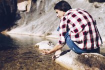 Visão traseira da mulher sentada na rocha e tocando água limpa no lago — Fotografia de Stock
