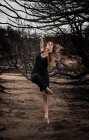 Jovem bailarina em desgaste preto com as mãos esticadas posando entre madeiras secas — Fotografia de Stock