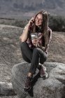 Smiling young woman in sunglasses with cup of drink talking on mobile phone and sitting on rock — Stock Photo