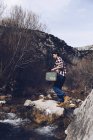 Seitenansicht einer lässigen Frau, die einen Koffer trägt und in der Natur auf Felsen von klarem Wasserstrom springt — Stockfoto